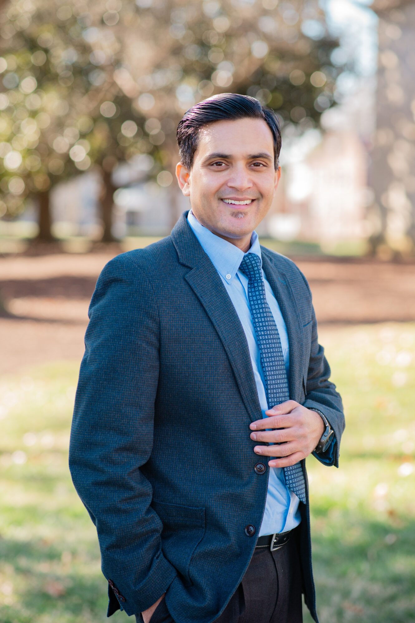 Man in a suit smiling outdoors with trees and grass in background.