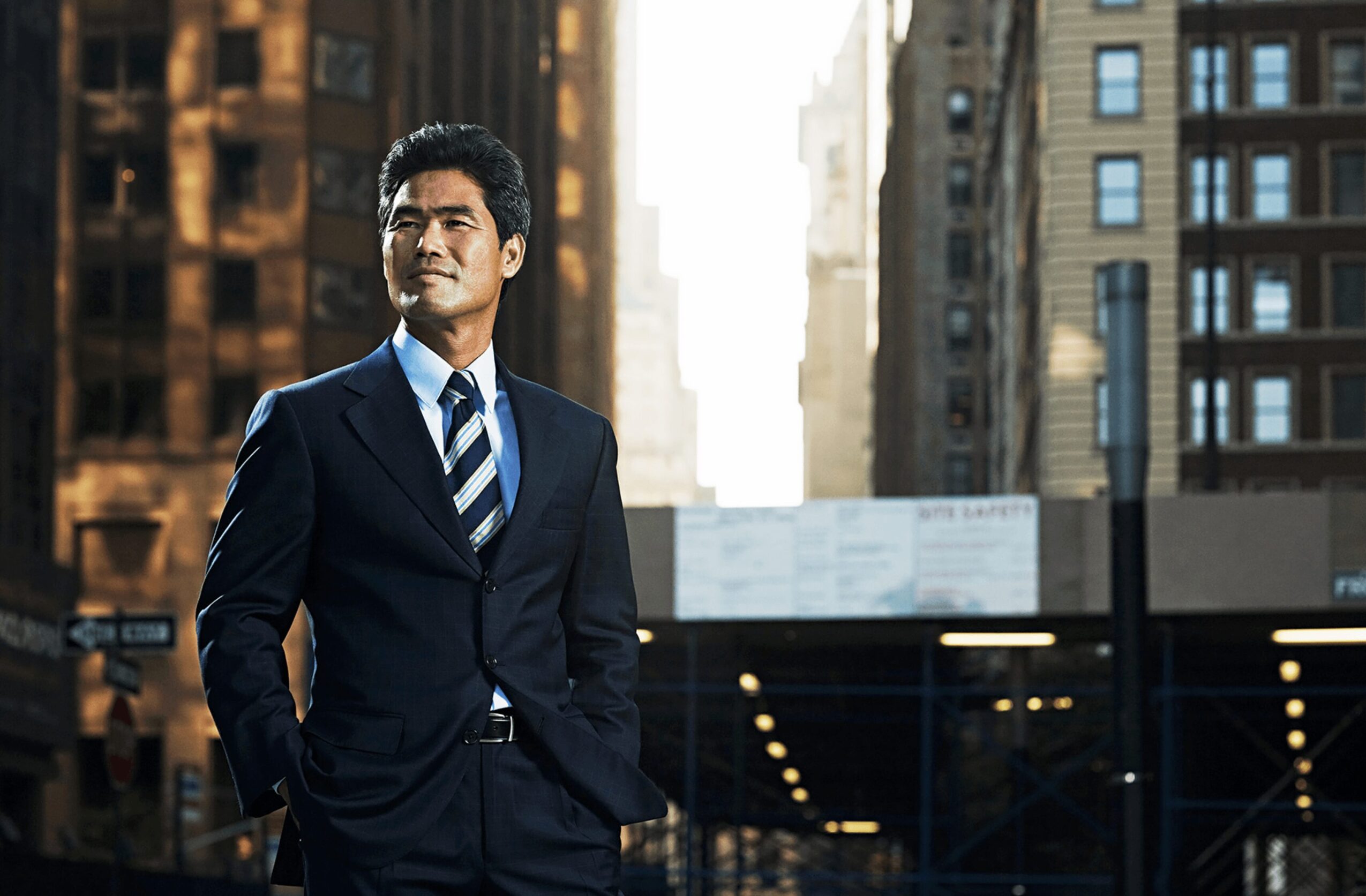 A man in a suit stands outdoors in a city with tall buildings, sunlight behind him, hands in pockets.