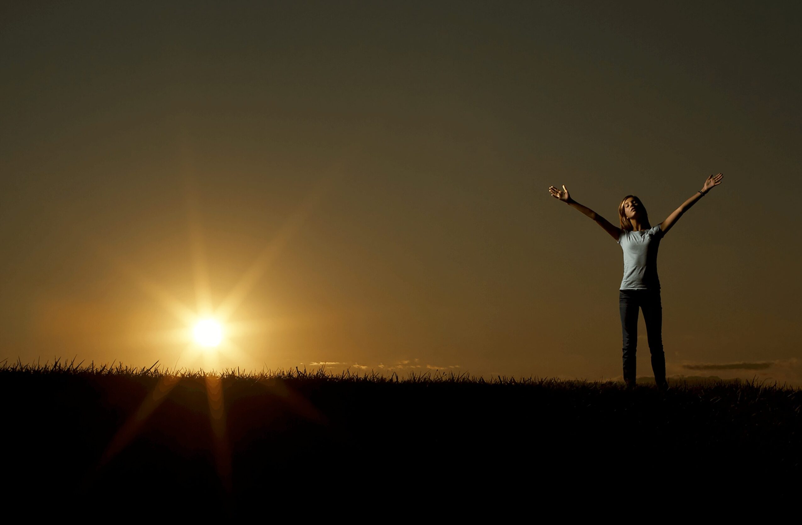 Person standing outdoors with arms raised at sunset, sun near horizon, dark sky, grassy ground.
