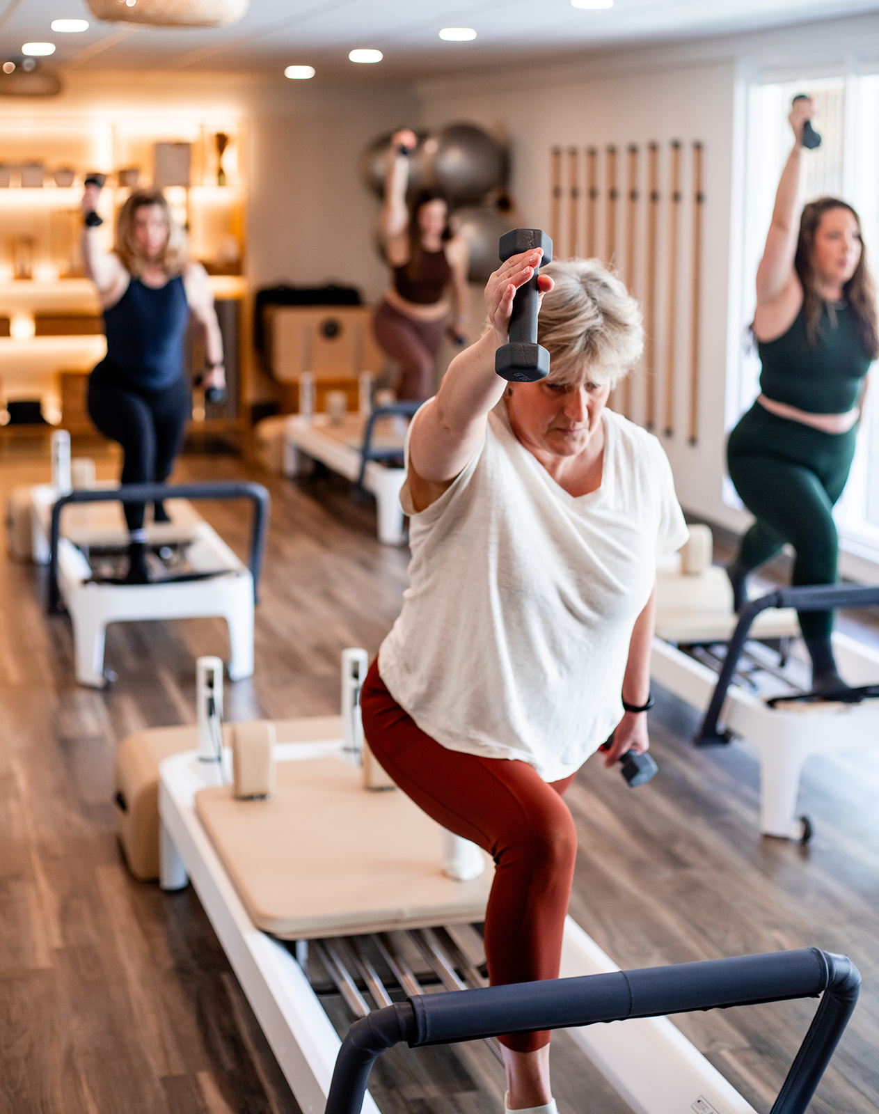 Group of women exercising with dumbbells and resistance bands in a fitness studio, some on Pilates reformers, in a well-lit room.