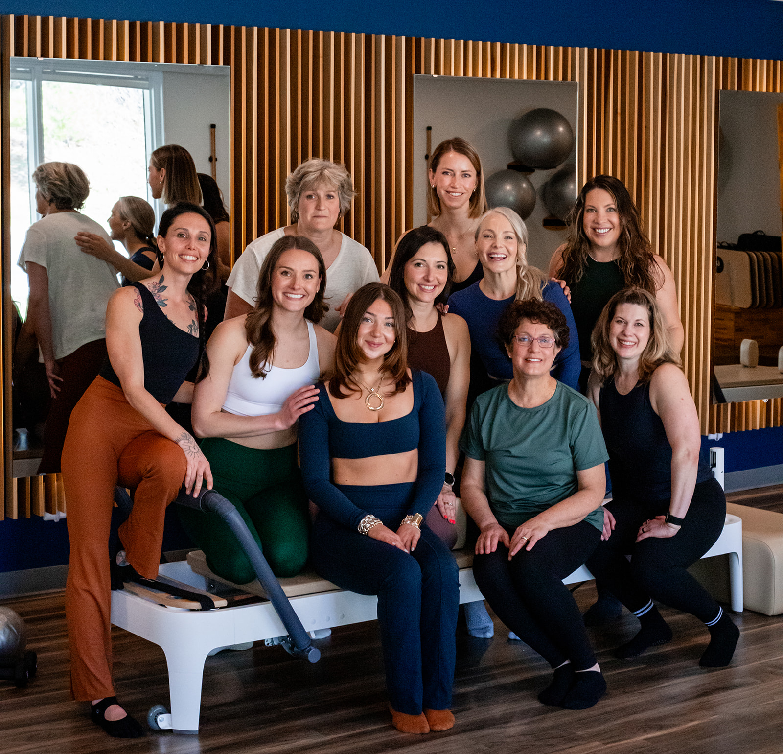 Group of ten women sitting and standing in a fitness studio, smiling, with wooden wall decor and exercise balls in the background.