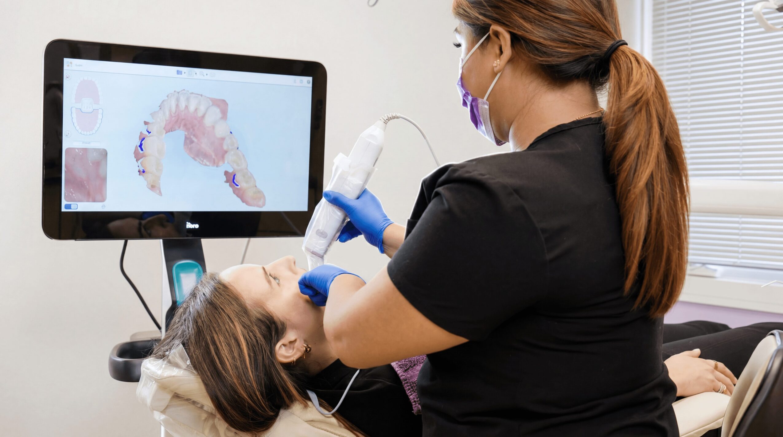 Dental professional using a device on a patient lying in a dental chair, with a monitor displaying a dental scan, in a clinic.