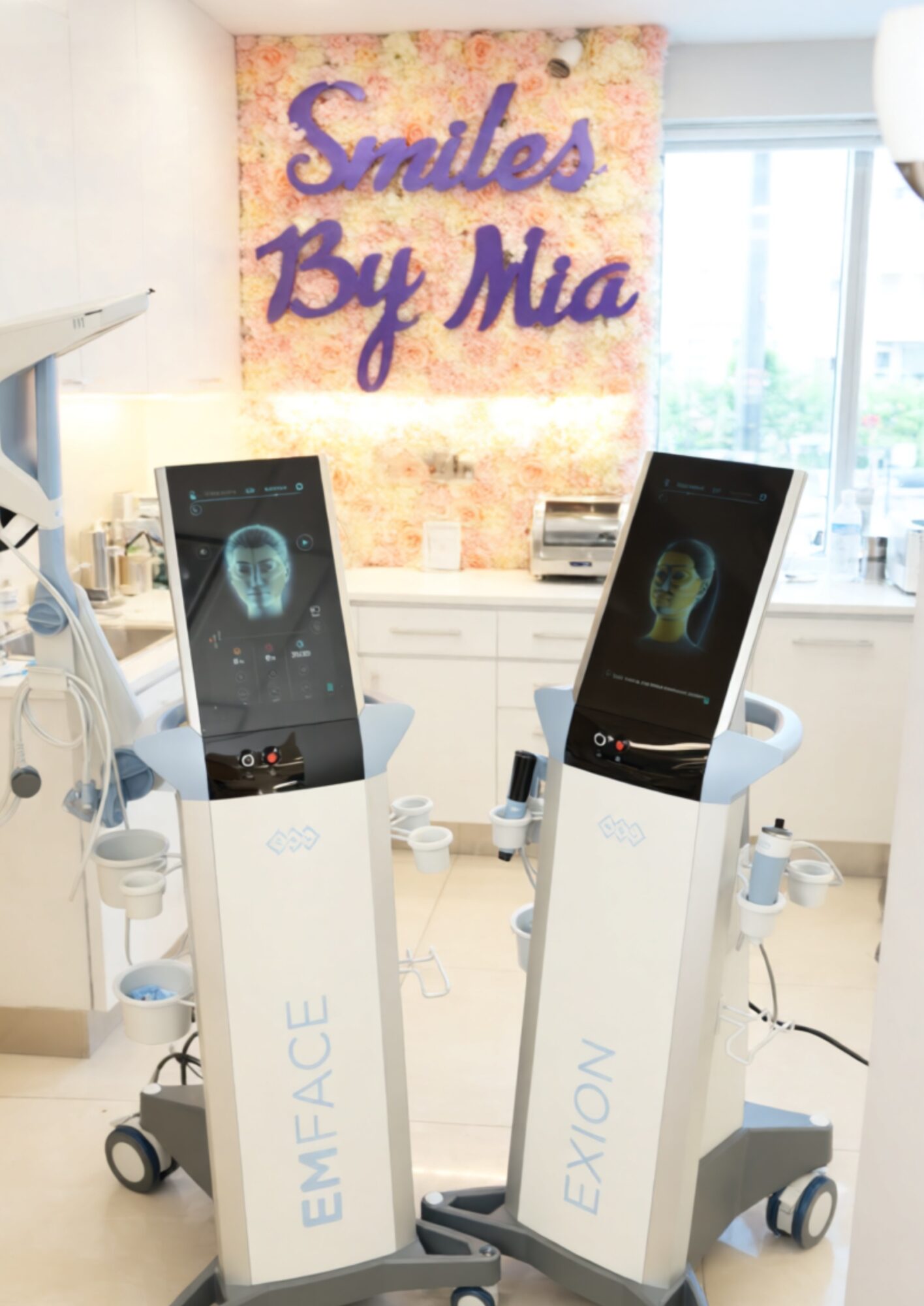 Two medical imaging machines with screens displaying brain scans in a bright clinic room, with a colorful sign reading 'Smiles By Mia' in the background.