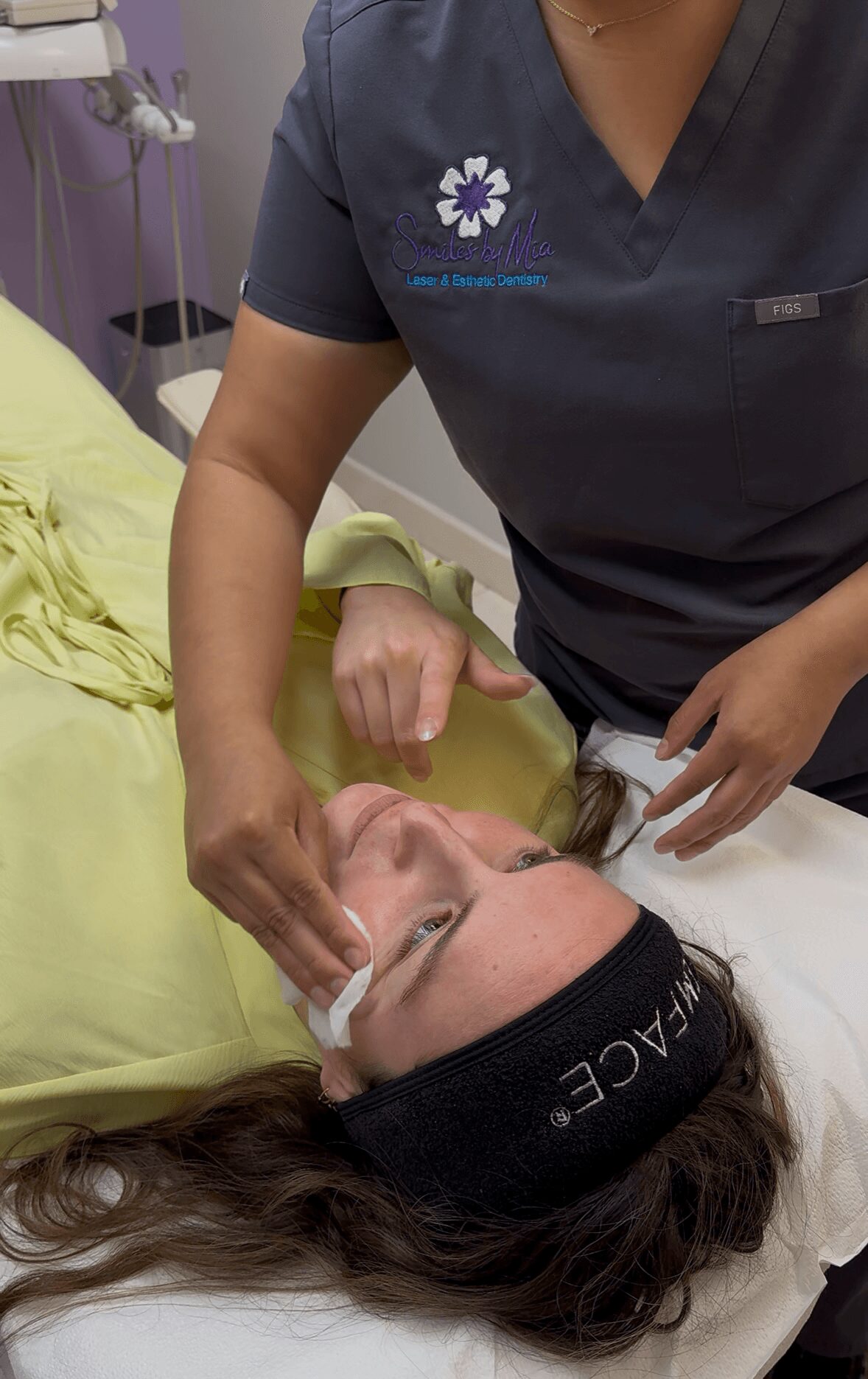 A woman lying on a bed with a headband, receiving facial treatment from a professional in scrubs.