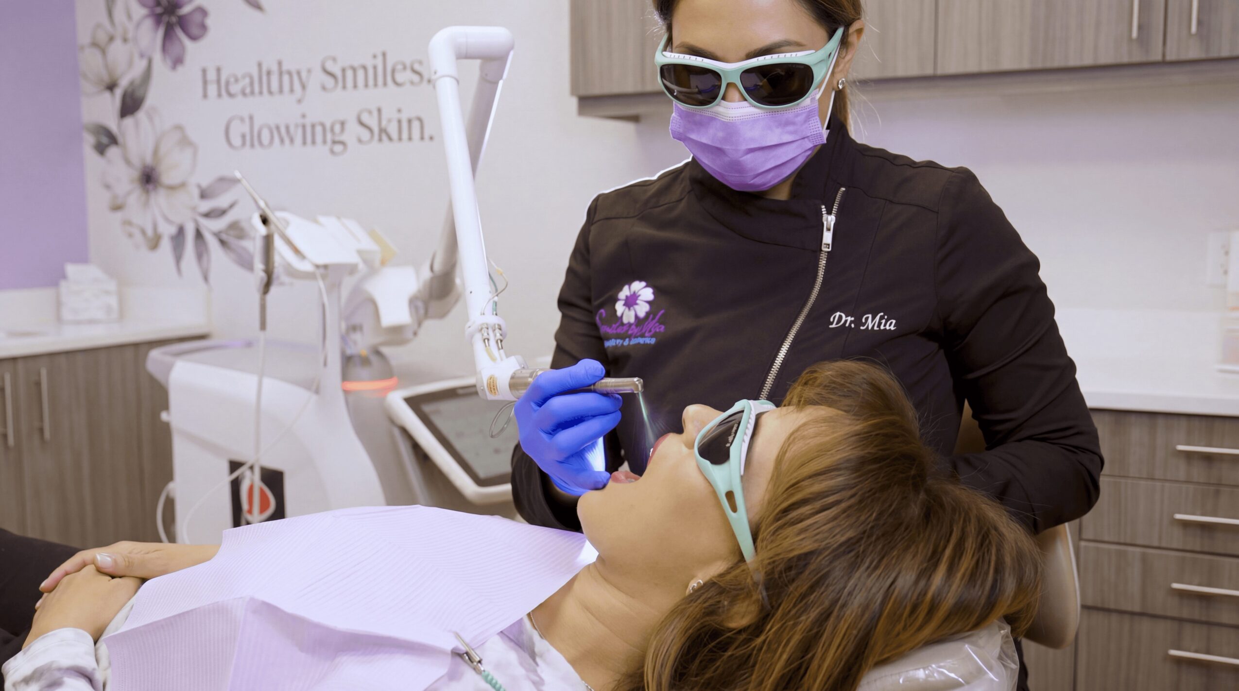 Dentist wearing purple mask and gloves examines patient lying in dental chair, patient wearing glasses, in a dental office.