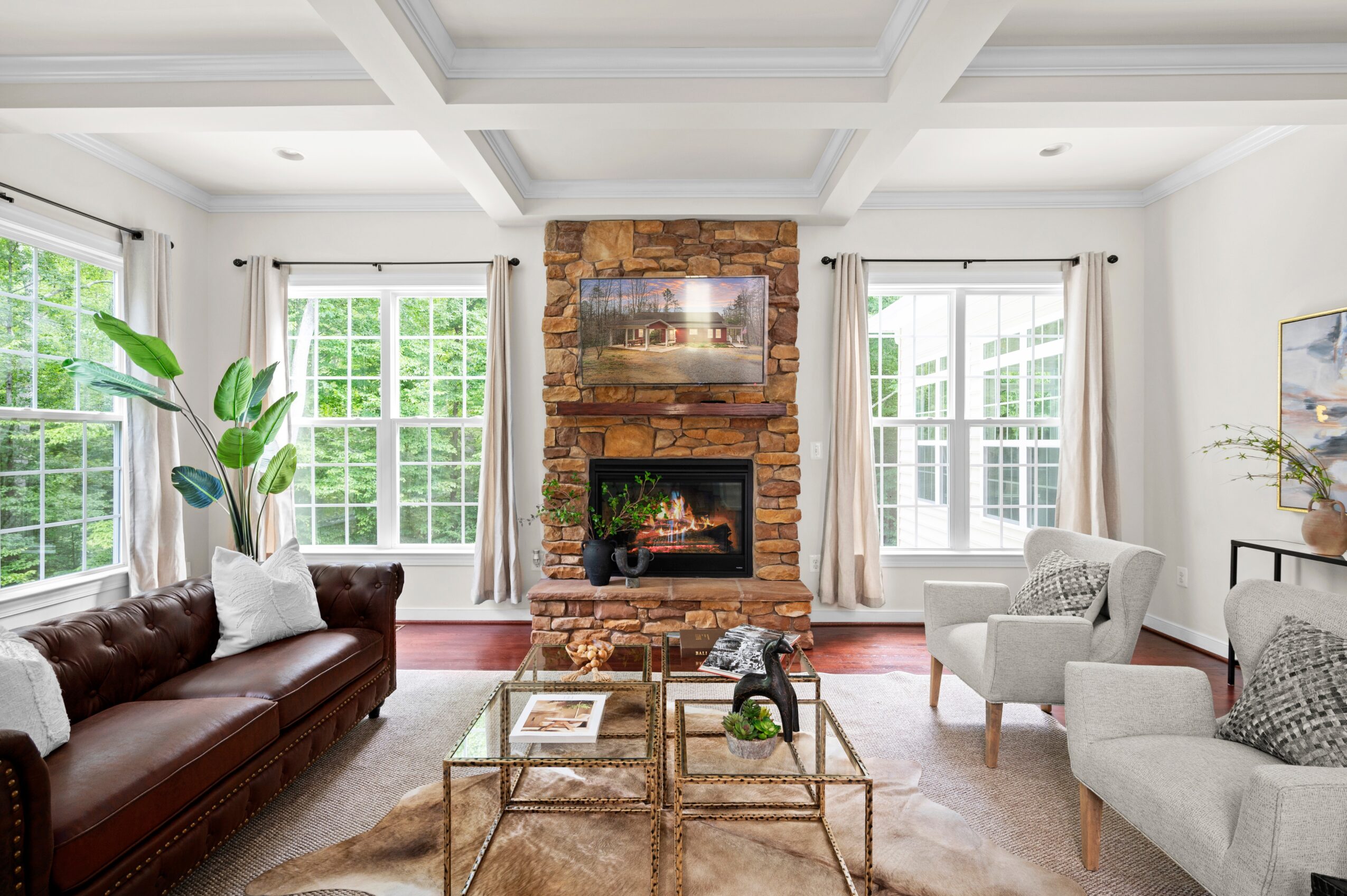 Living room with brick fireplace, large windows, white walls, and furniture including a brown sofa and white armchairs.