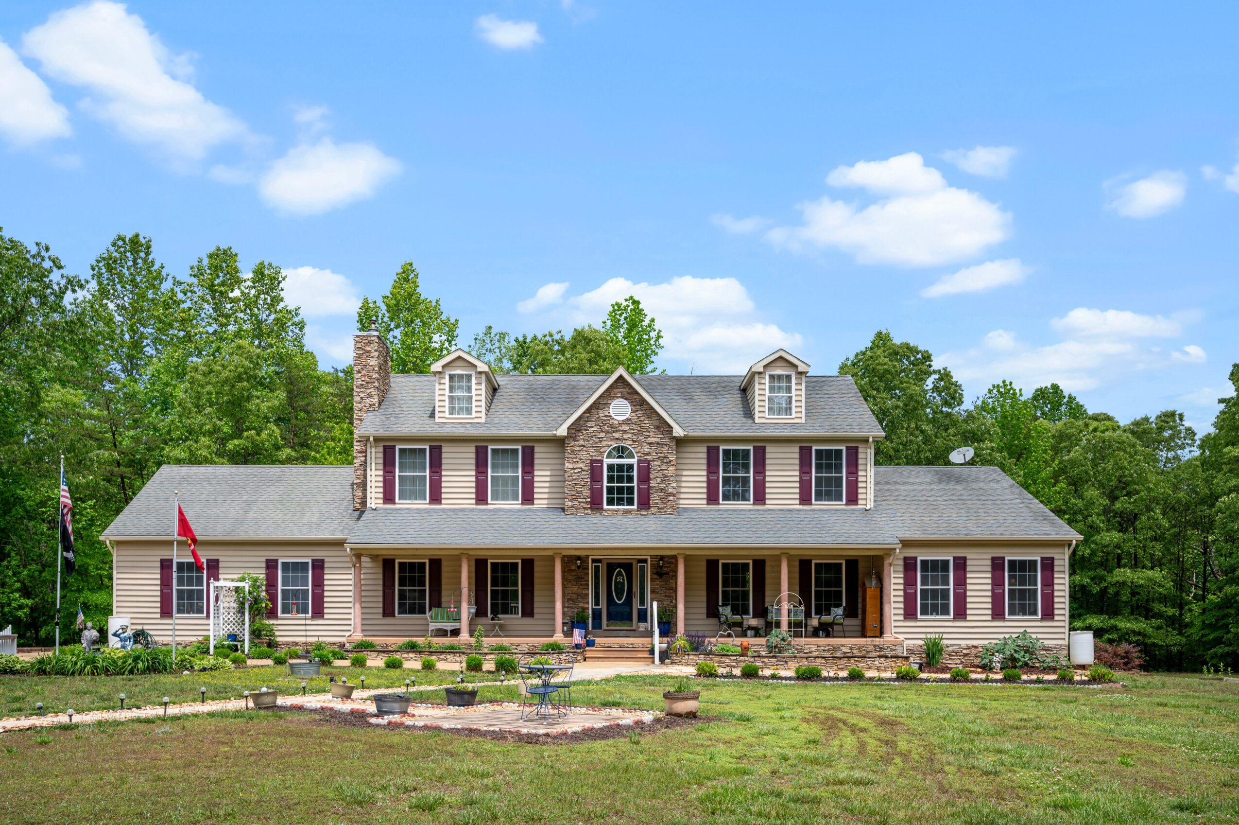 Large two-story house with a front porch, surrounded by trees and a lawn, under a blue sky with clouds.