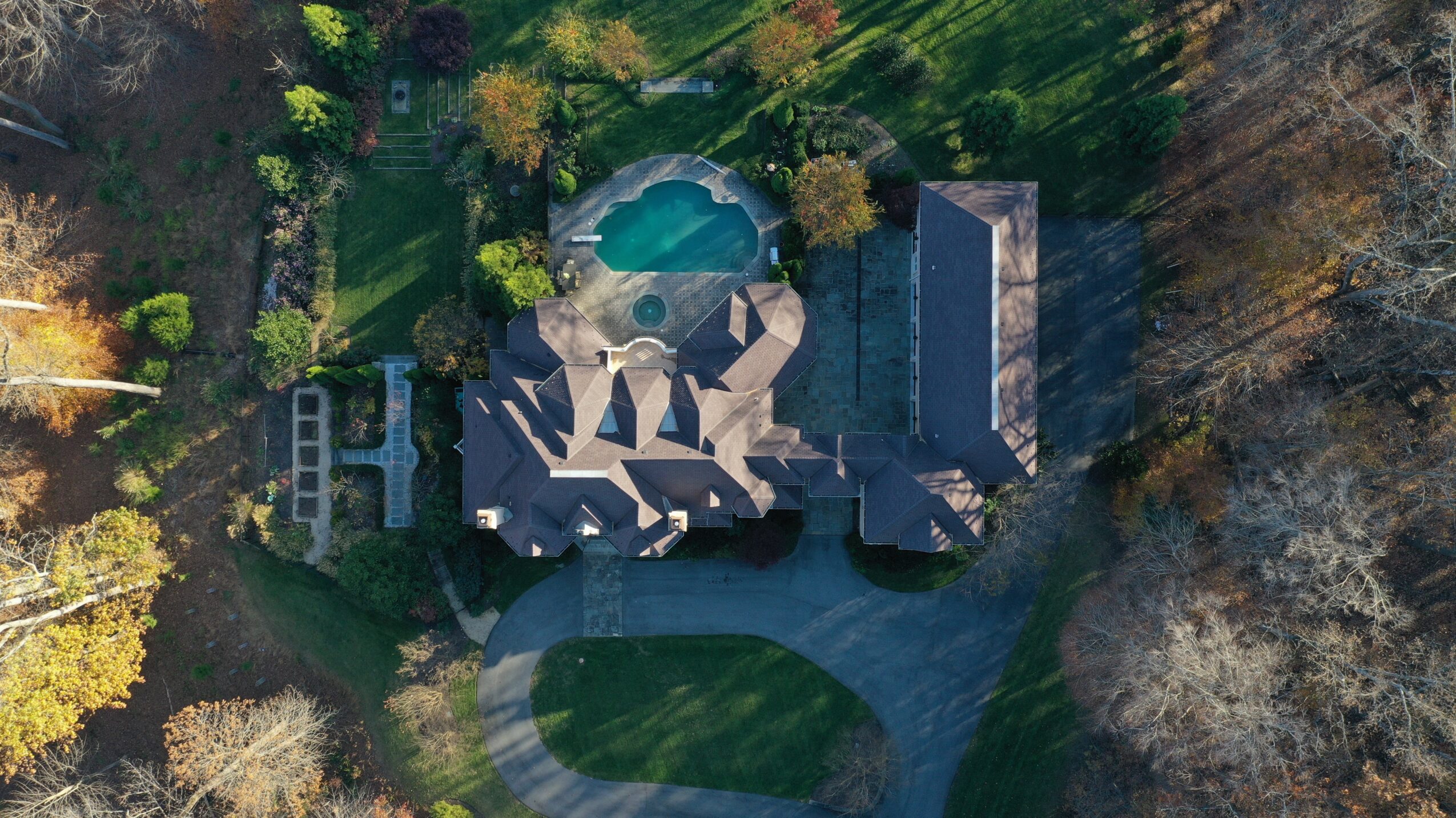 Aerial view of a large house with a swimming pool, surrounded by trees and a driveway, on a sunny day.