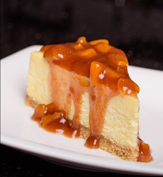 Slice of cheesecake with caramel sauce and chunks on top on a white plate, dark background.