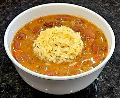 Bowl of soup with rice in the center, containing vegetables and meat, on a dark surface.