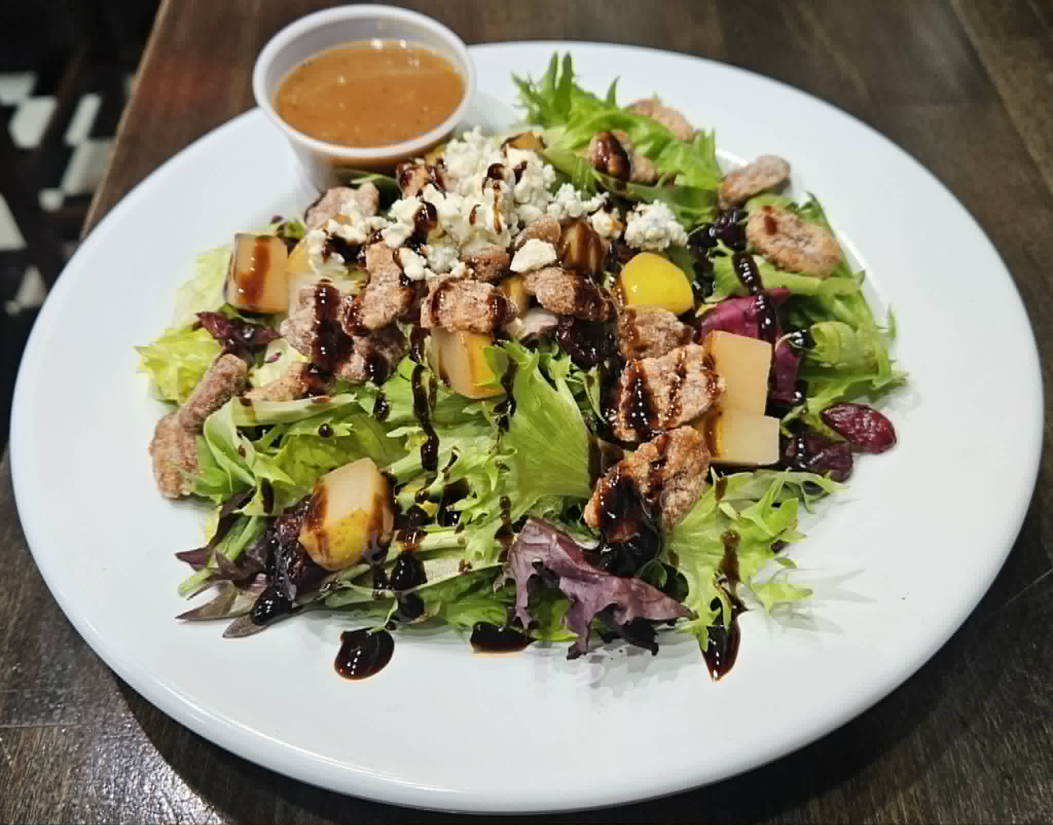 Mixed green salad with grilled chicken, cheese, fruit, and a side of dressing in a small bowl, on a white plate.