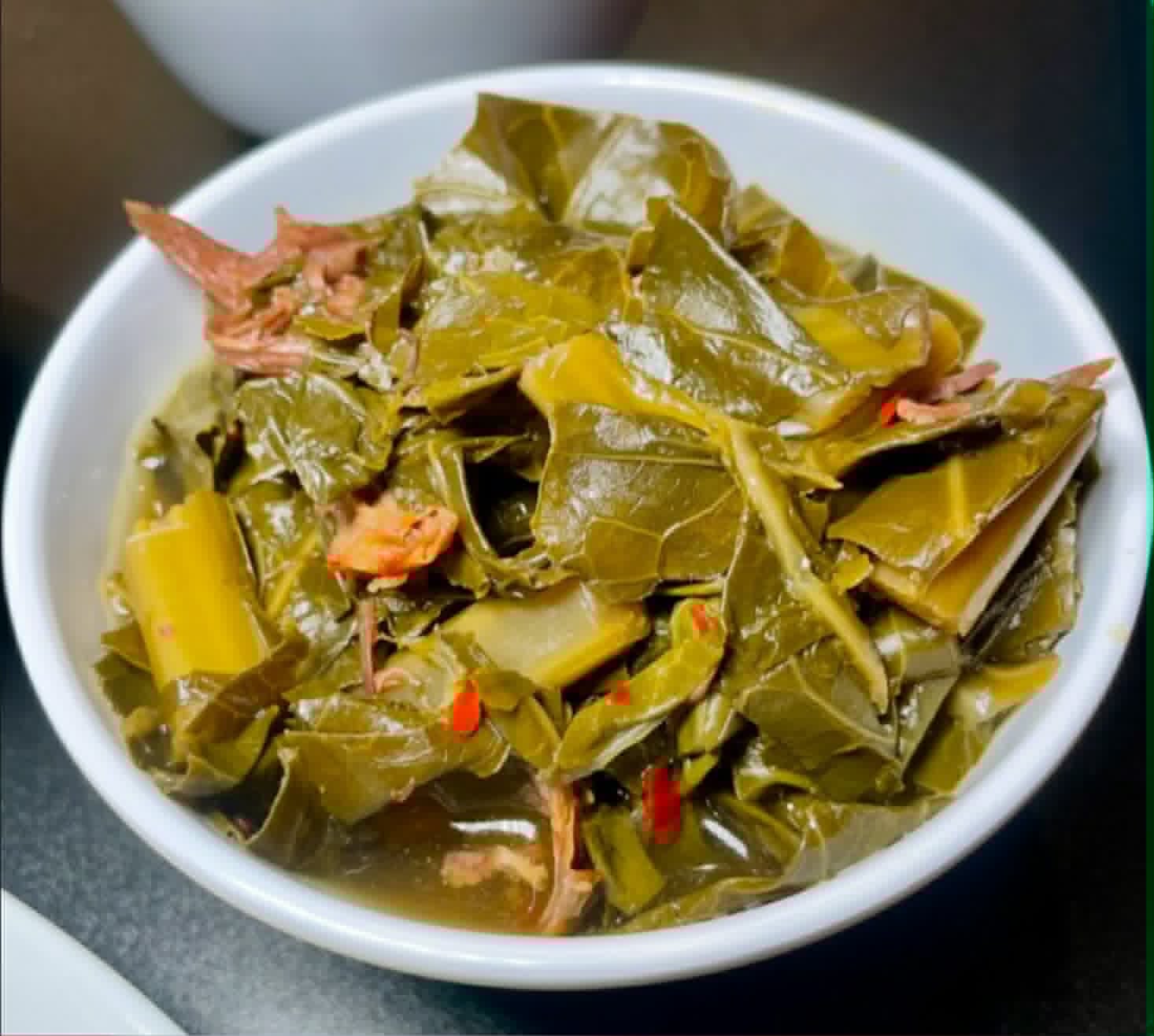 Bowl of cooked leafy greens with small pieces of meat and red chili flakes, on a dark surface.