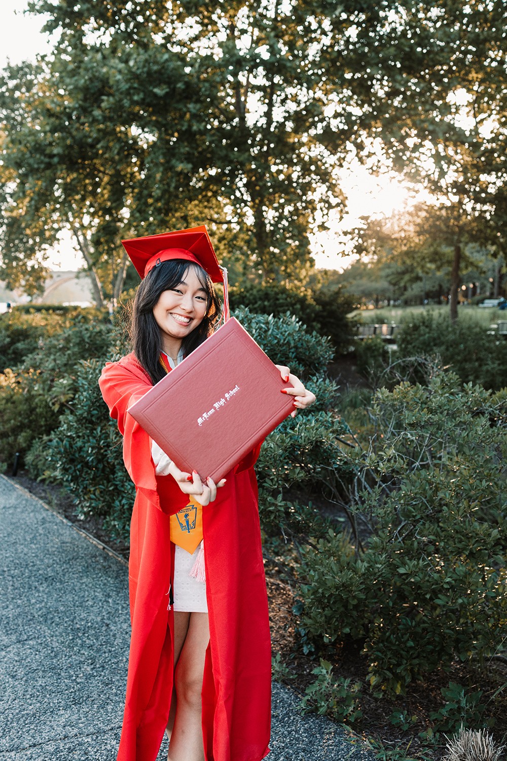Young woman in graduation gown and cap holding a diploma outdoors with trees and sunlight in background.