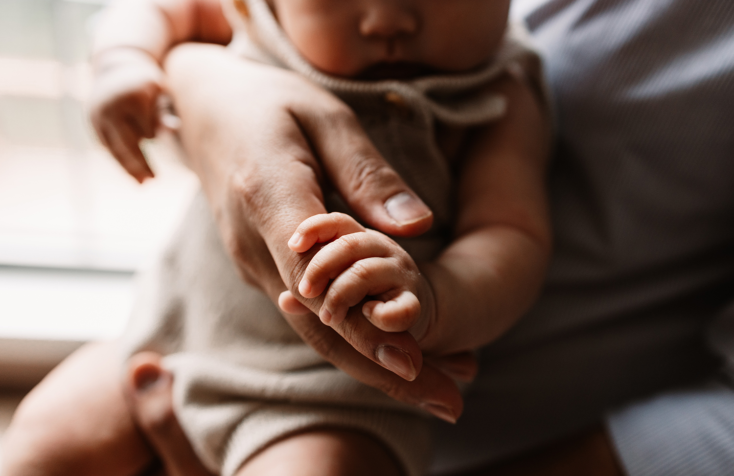 Adult holding a baby with both hands, baby’s hand gripping adult’s finger, soft natural lighting, close-up shot.