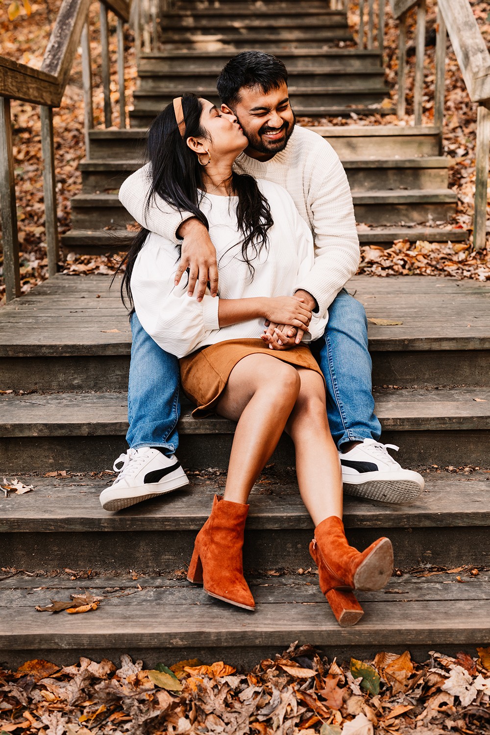 A smiling couple sitting on outdoor wooden stairs surrounded by autumn leaves, embracing each other, woman wearing orange boots, man in white sweatshirt.