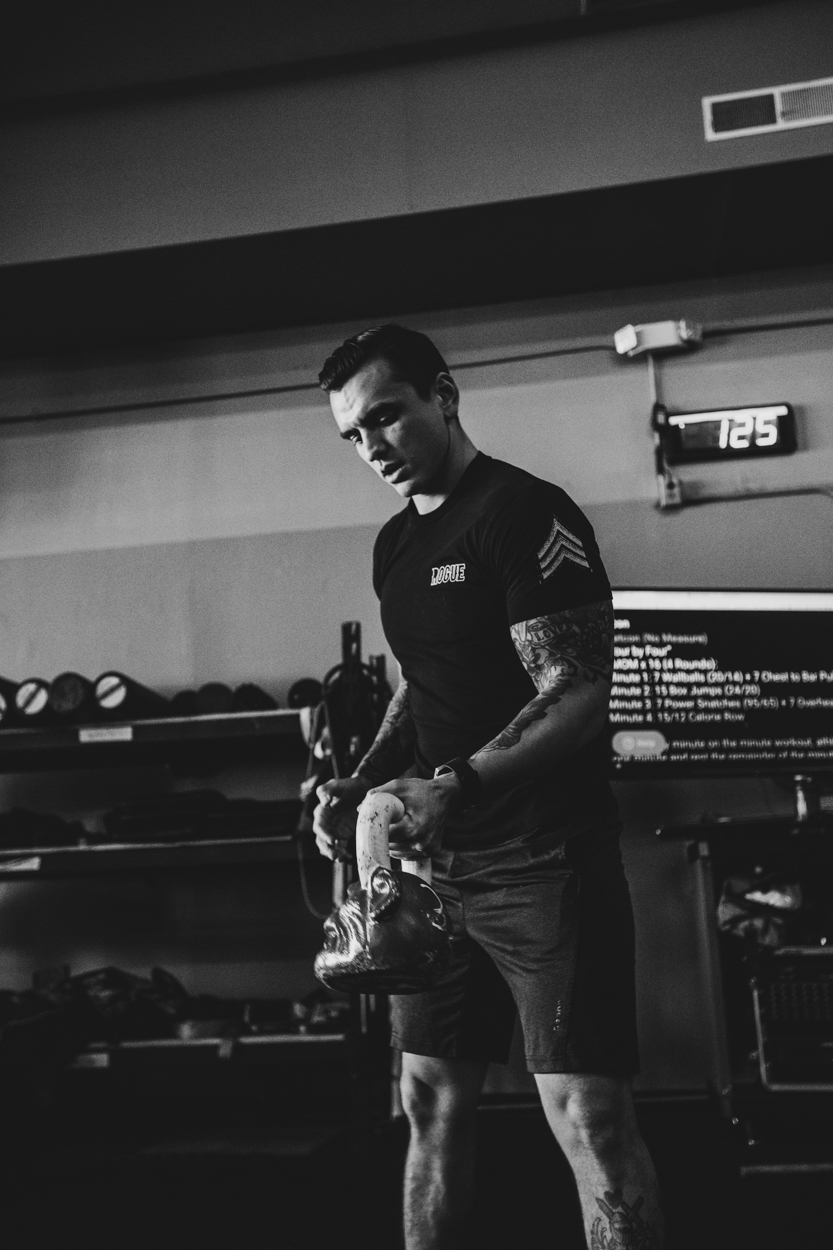 Man with tattoos holding a kettlebell in a gym, shelves with equipment in background