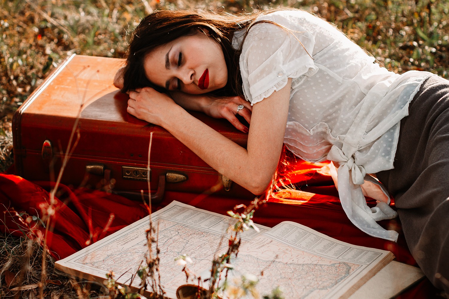 Woman lying on grass with a red suitcase and open book, resting her head on her arm, outdoors.