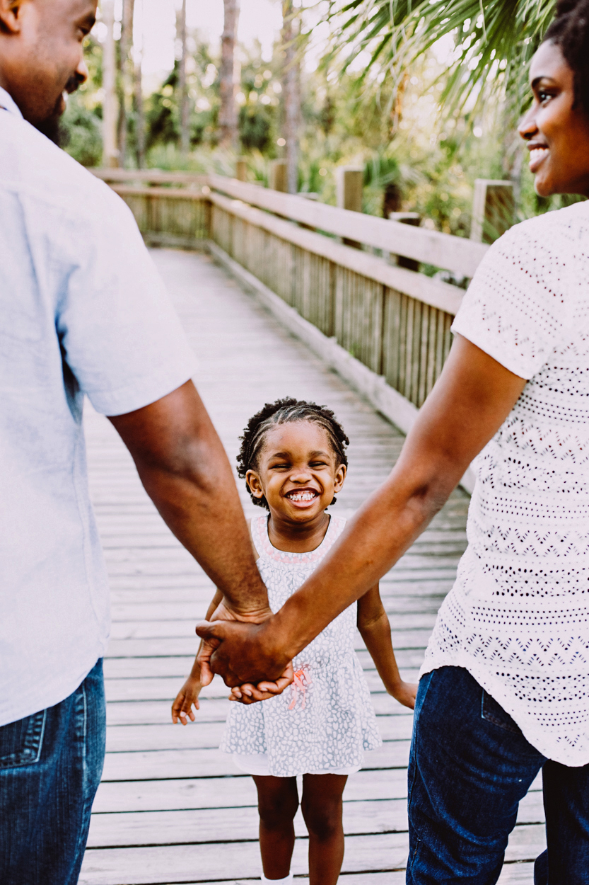 A family holding hands with a smiling child on a wooden bridge outdoors, surrounded by greenery.
