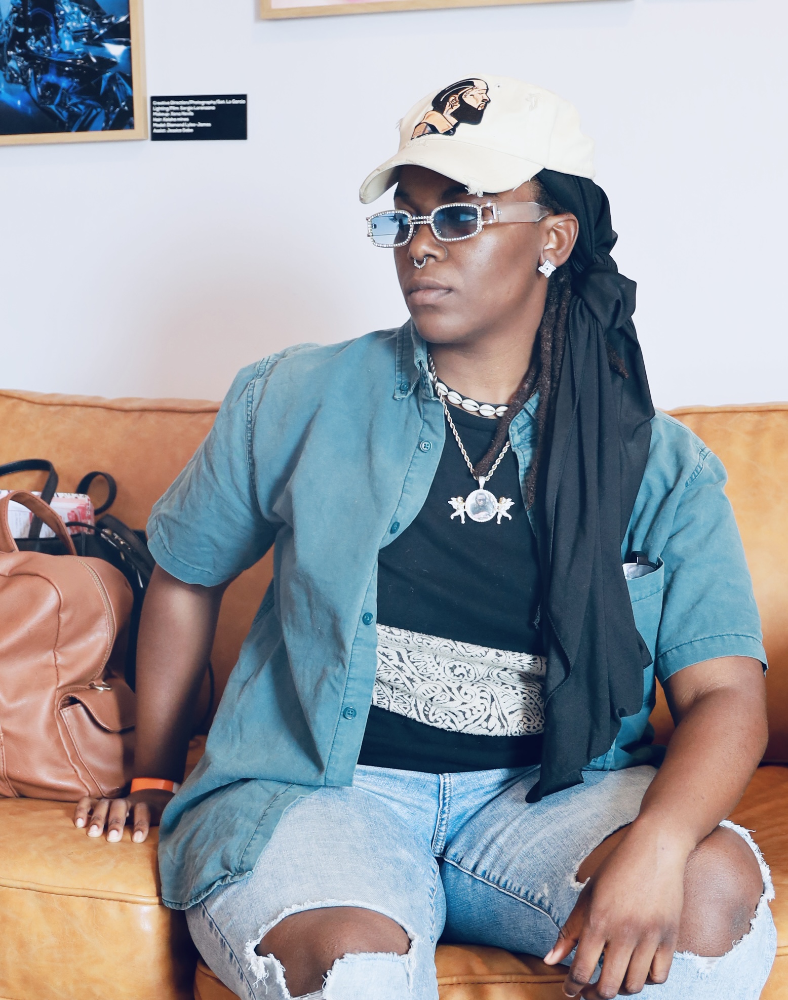 Woman sitting on a couch wearing sunglasses, a cap, and layered clothing, with a bag beside her.