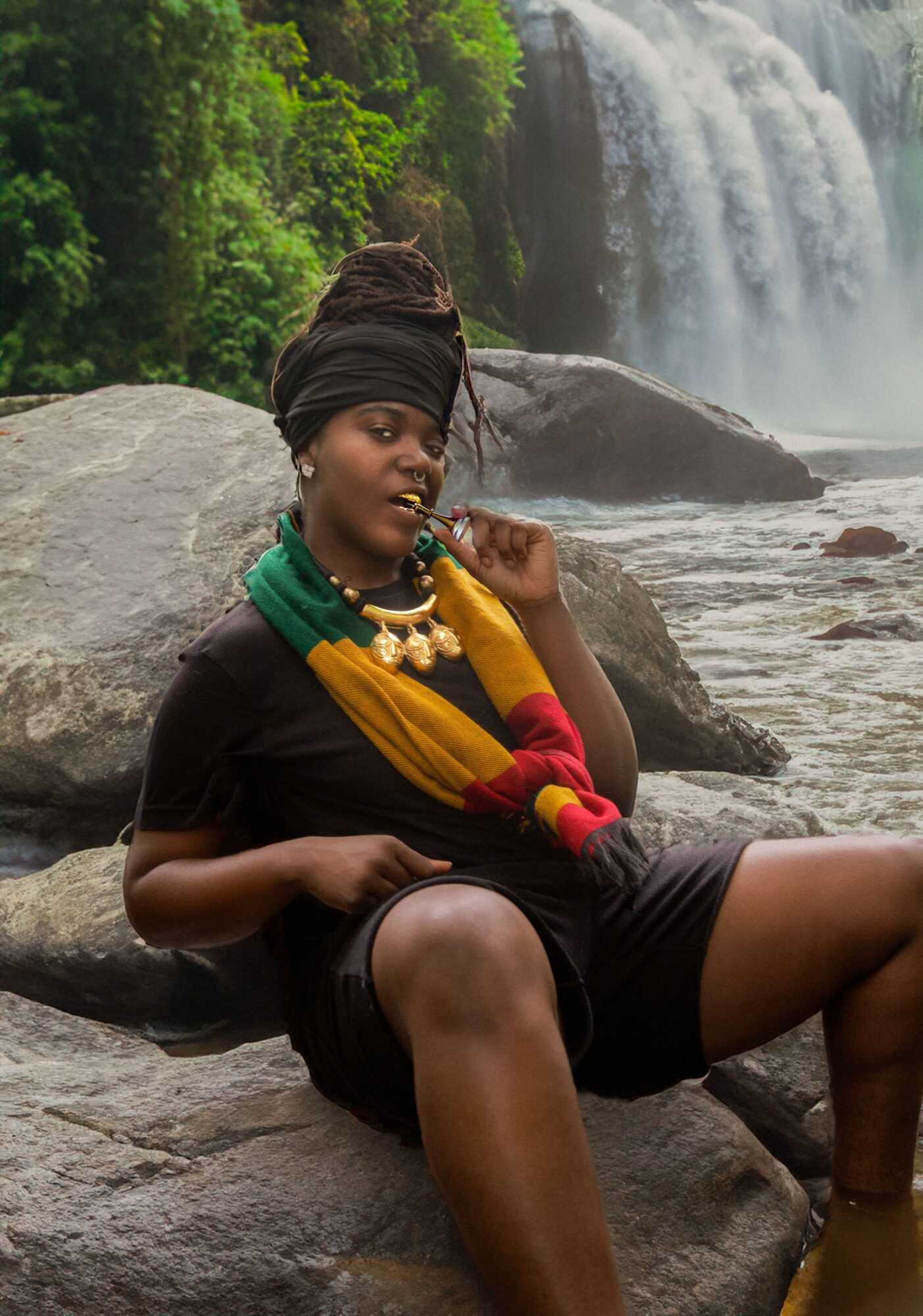 Woman sitting on rocks near a waterfall, wearing a black dress, colorful scarf, and jewelry, with lush greenery in background.