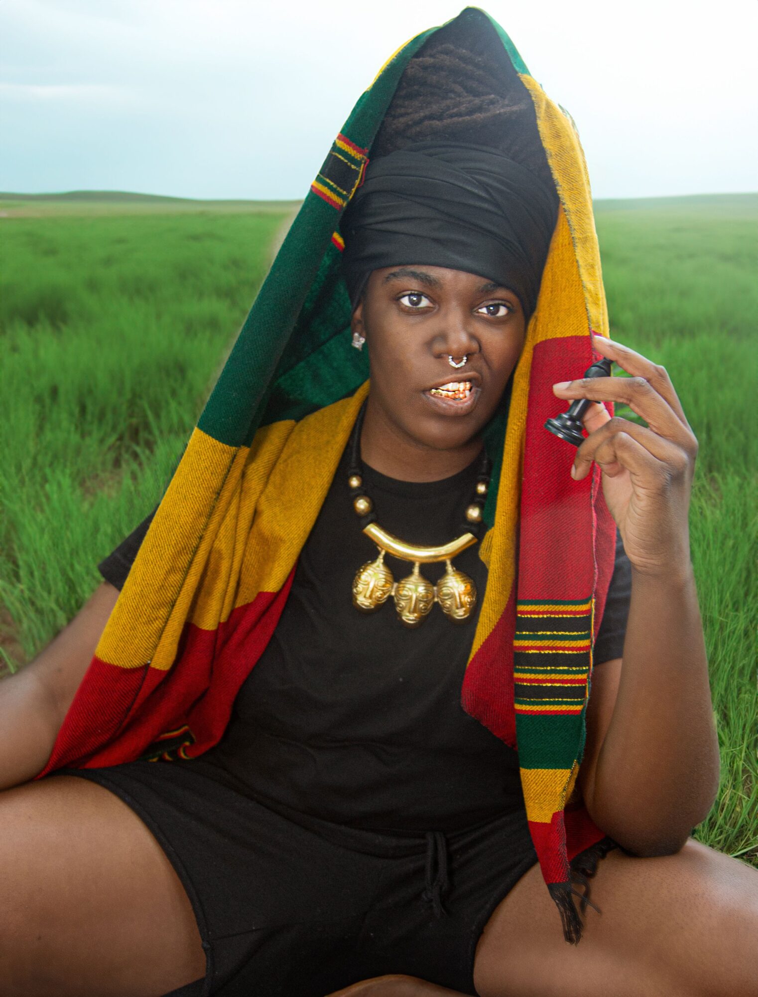 Woman with traditional headwrap and jewelry sitting outdoors in a grassy field, holding a finger to her face.