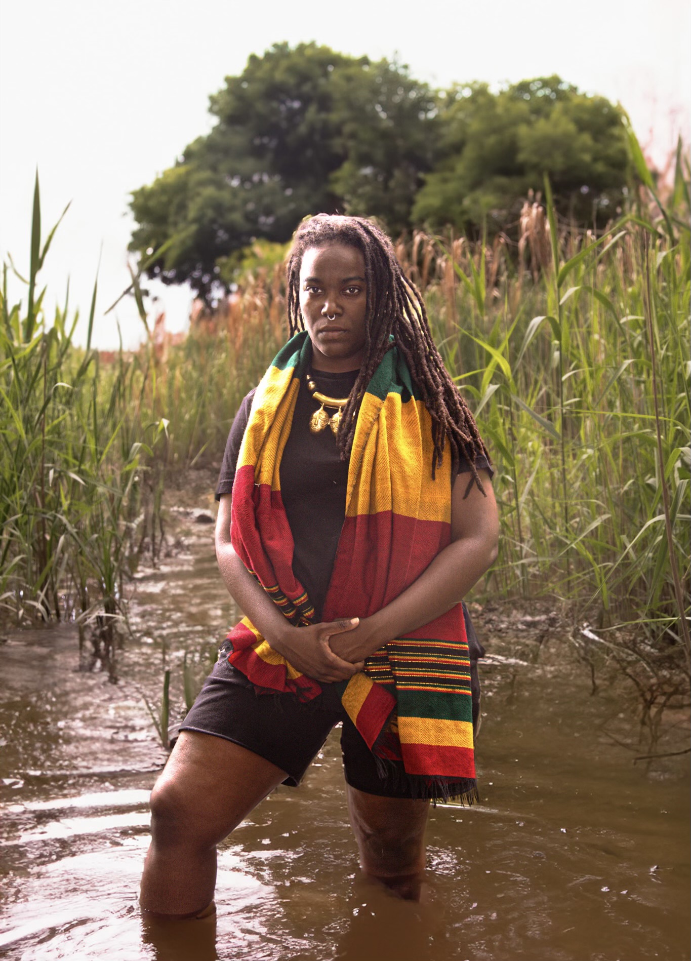 Woman standing in water with tall grass and trees in background, wearing a colorful scarf and black outfit.