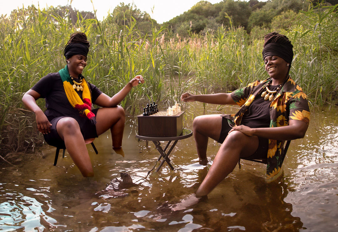 Two women sit in water with chairs, smiling, surrounded by tall grass and trees, playing a game with small objects on a table.