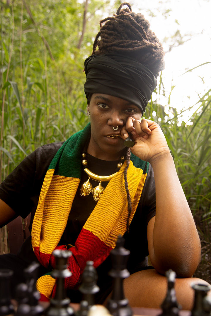 Woman with dreadlocks wearing a headwrap, colorful scarf, and jewelry, sitting outdoors near greenery, looking at camera.