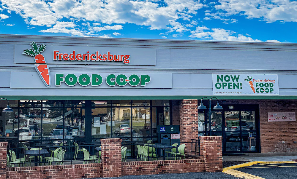 Facade of Food Coop store with signs, large windows, and brick columns, under a partly cloudy sky.