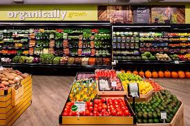 Grocery store produce section with fruits and vegetables on display, shelves in the background, and a wooden floor.