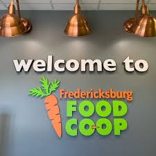 Sign with text 'welcome to Fredericksburg FOOD COOP' and a carrot graphic on a gray wall, illuminated by copper pendant lights.