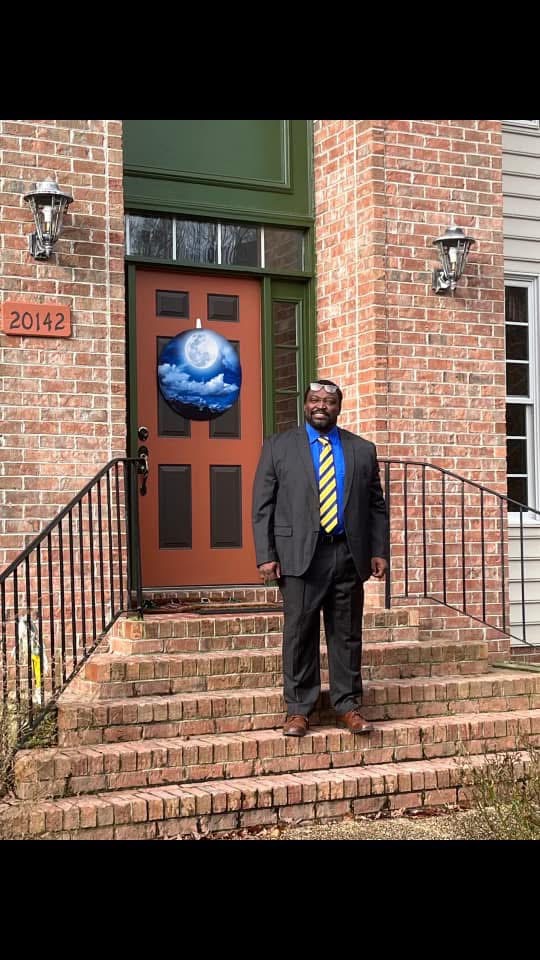 Man in suit with yellow striped tie standing on brick steps outside building with brick walls and green door.