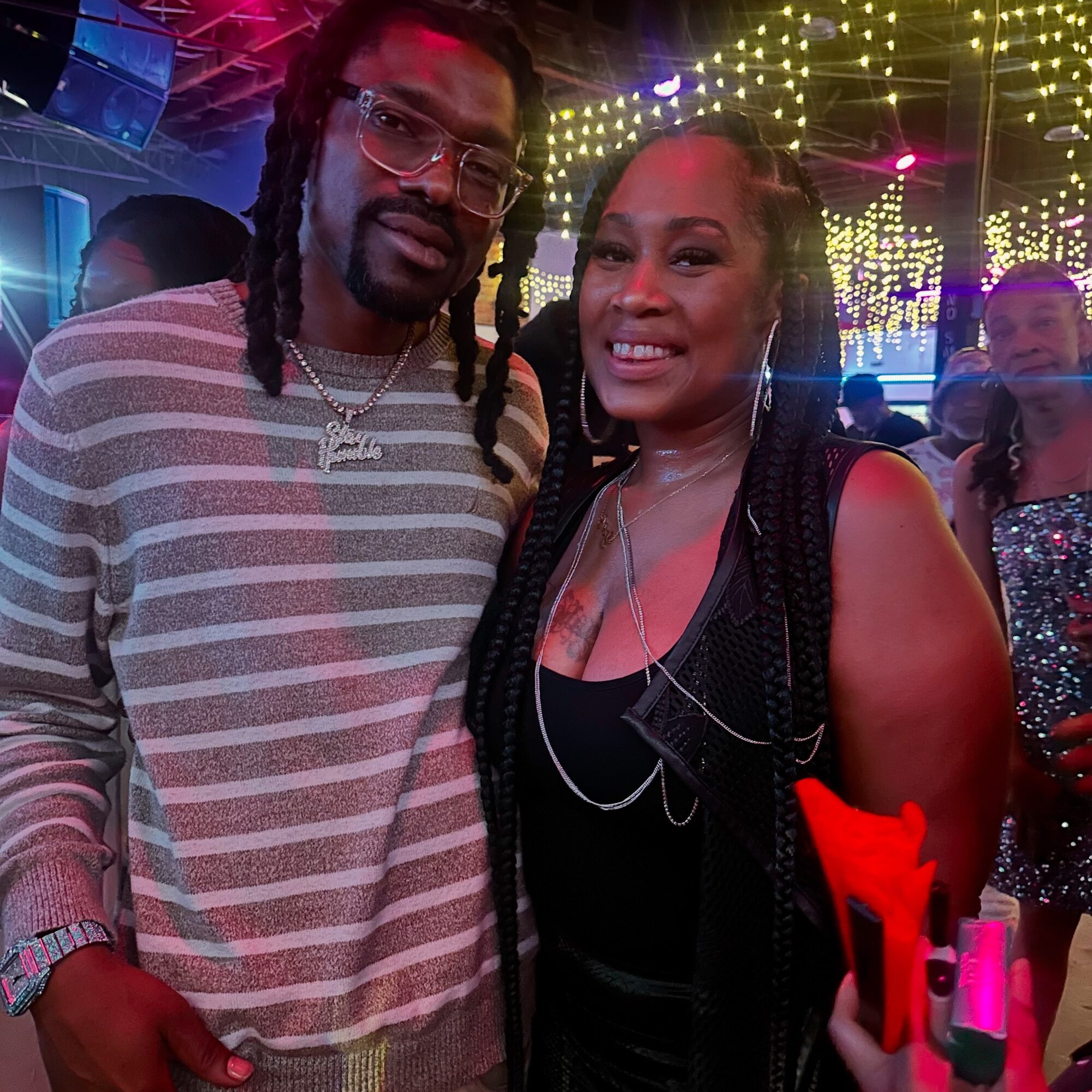 Two people smiling at a party with colorful lights and other guests in the background.