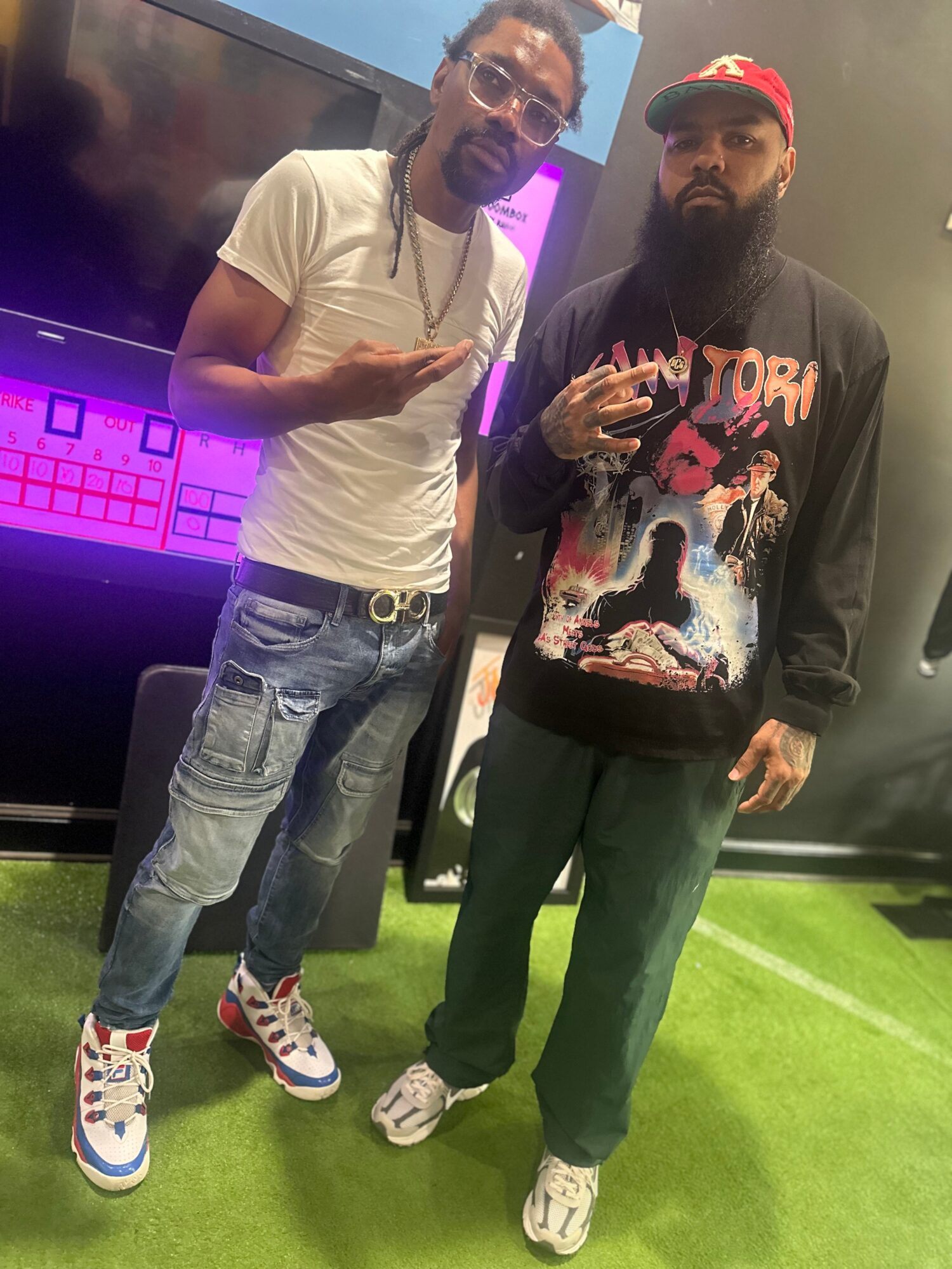 Two men stand indoors on a green floor, one wearing a white t-shirt and jeans, the other in a black hoodie and cap.