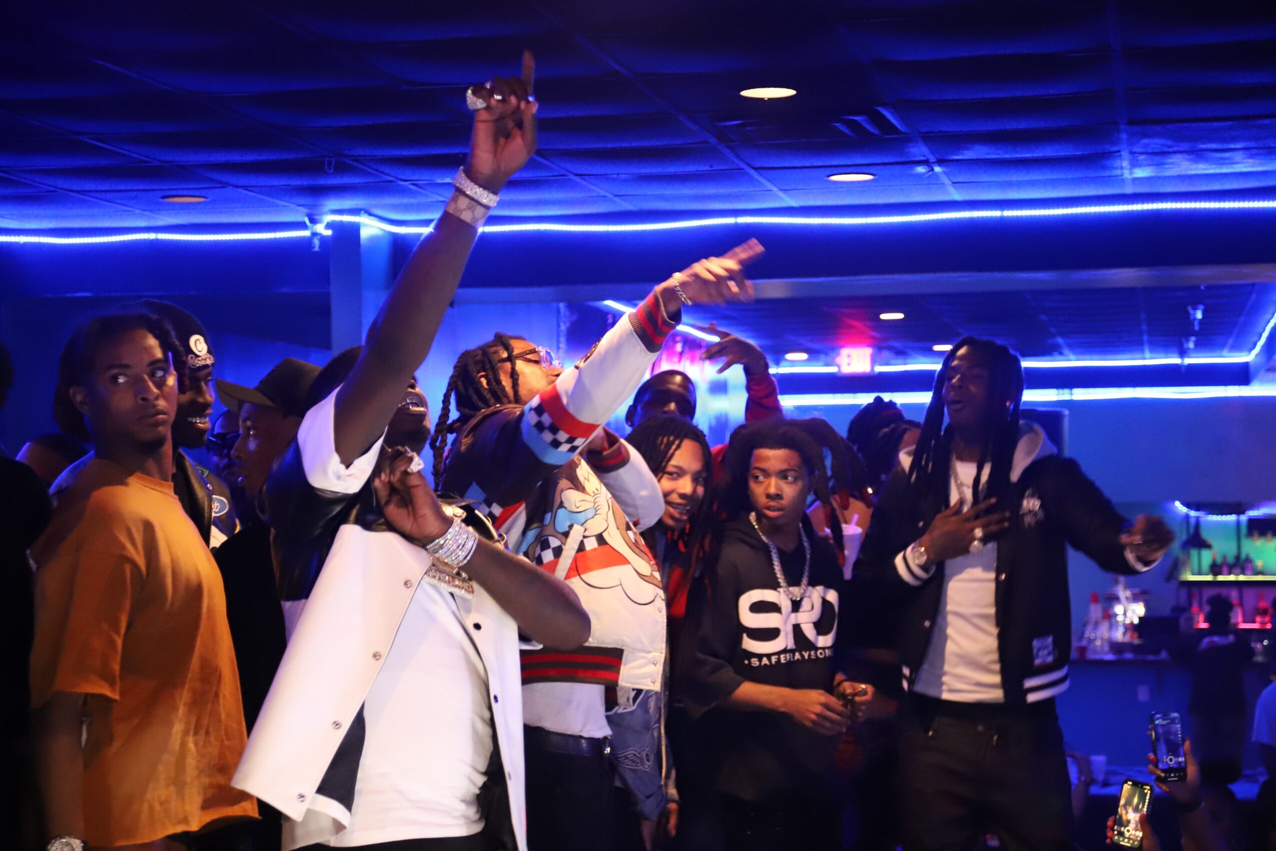 Group of people dancing in a nightclub with blue lighting, some raising hands, wearing casual clothing.