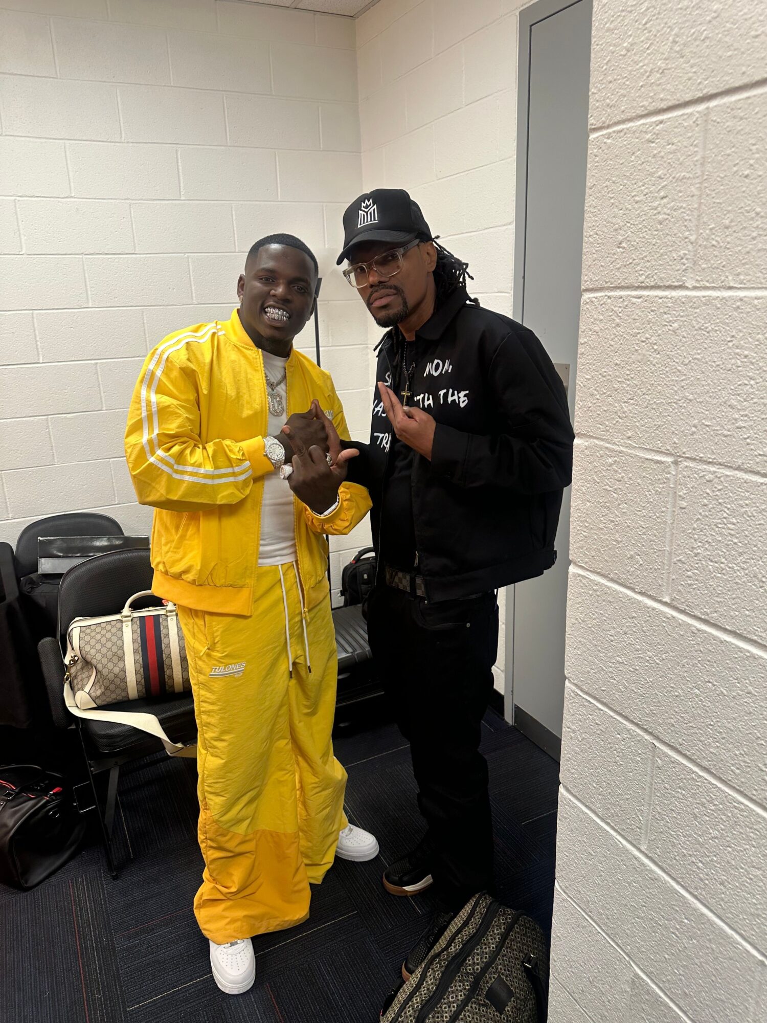 Two people standing indoors, one in a yellow outfit and the other in dark clothing, with luggage and a white wall background.