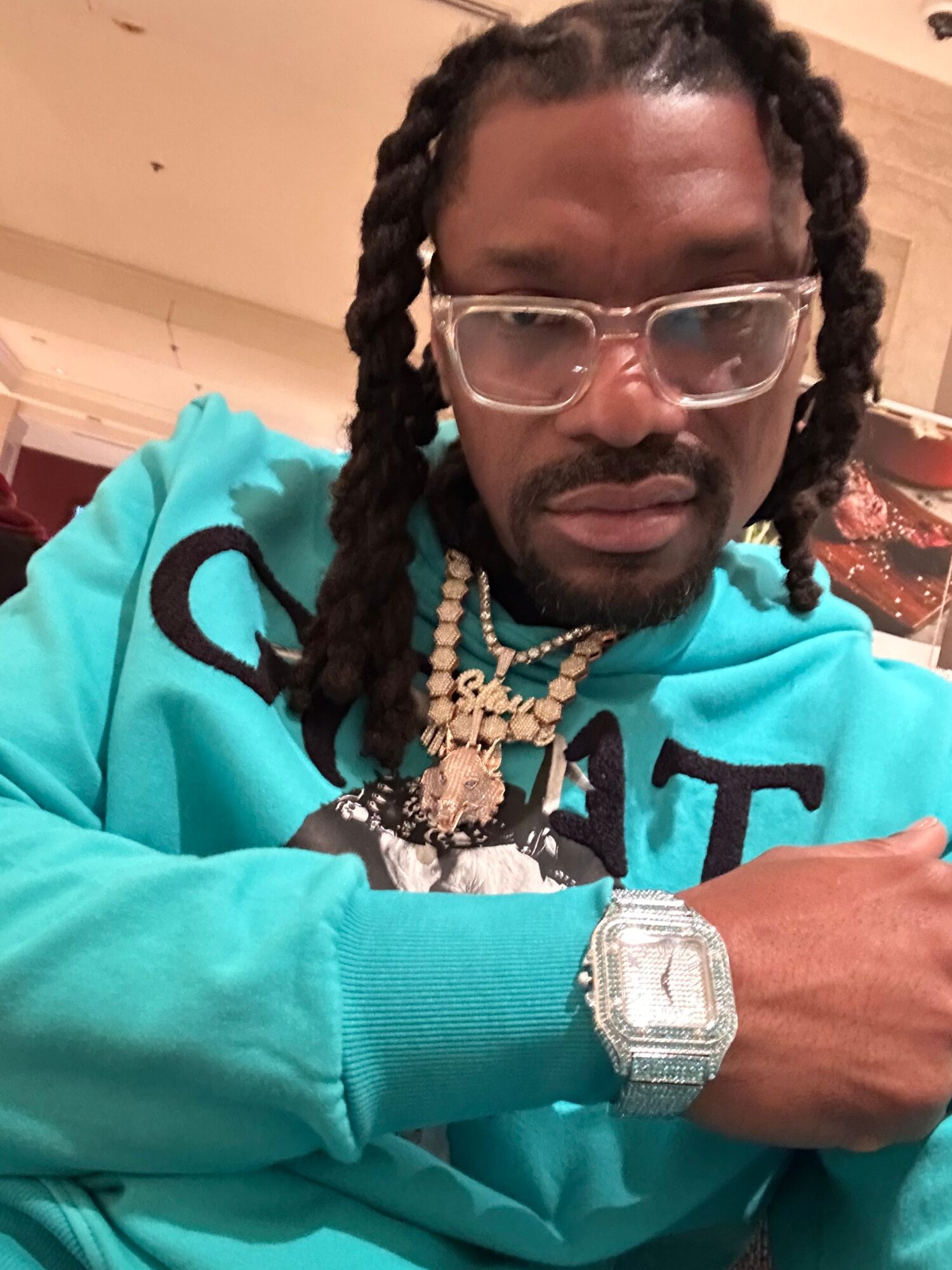 Man with glasses, braided hair, and jewelry wearing a turquoise hoodie, resting his arm on a surface.