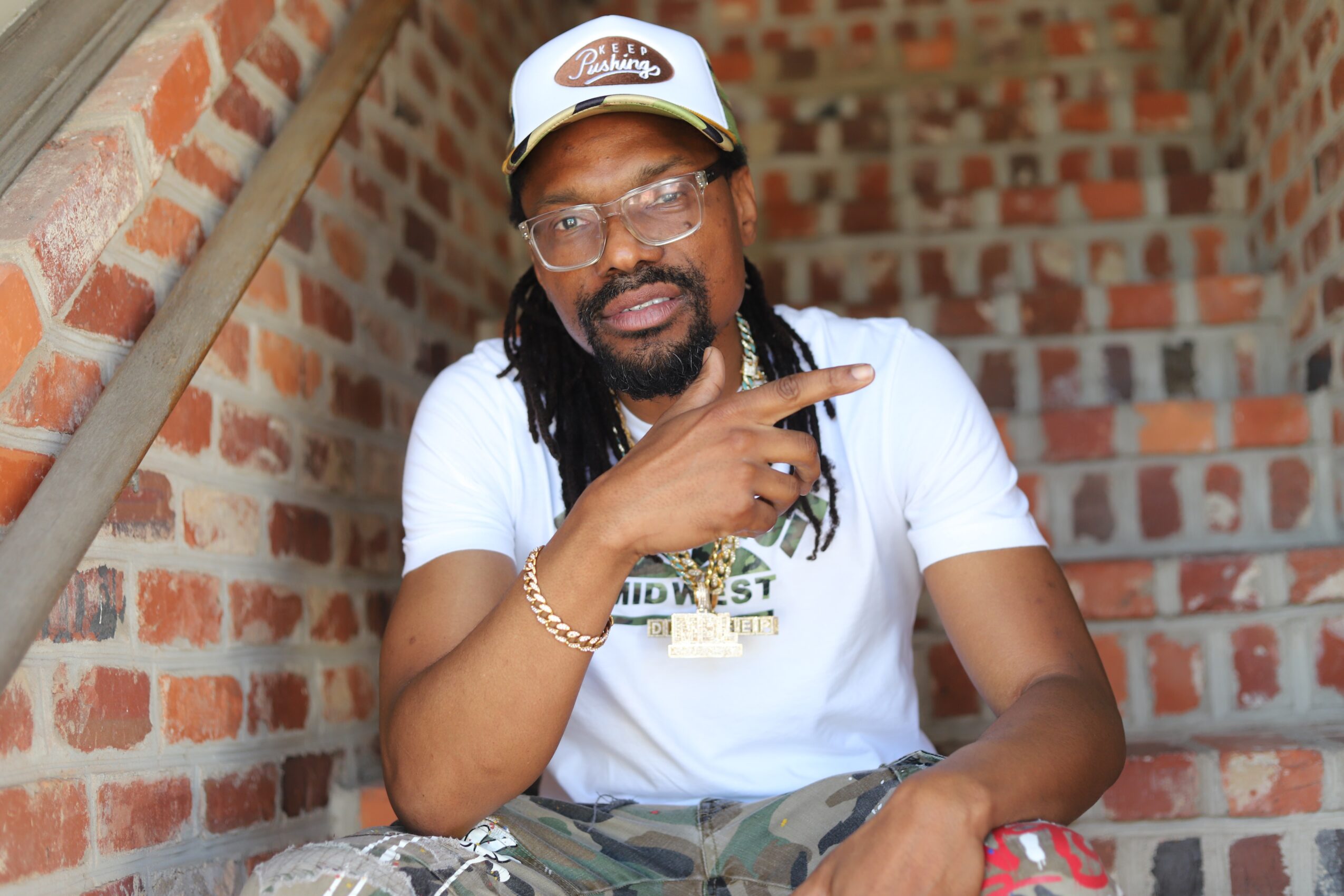 Man with glasses and a cap sitting on brick stairs, pointing with his right hand, wearing a white t-shirt and camouflage pants.