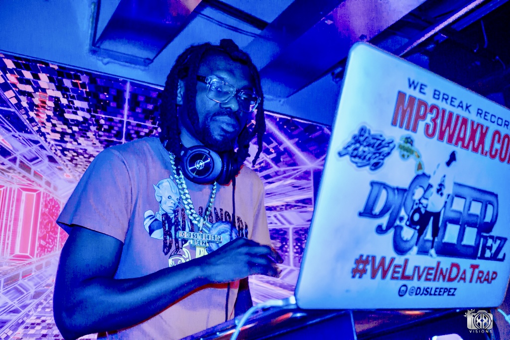 DJ with headphones in a colorful, illuminated setting, working at a laptop with stickers and text, including MP3WAXX.COM.
