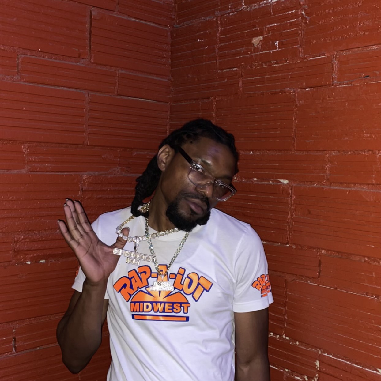 Man with glasses and dreadlocks wearing a white T-shirt with orange and blue text, standing against a red brick wall.