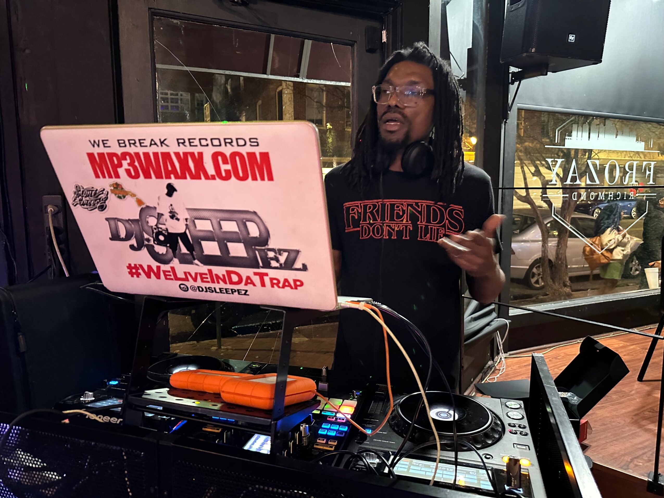 Person with long hair and glasses standing behind DJ equipment inside a venue, holding a phone, with a window showing outside scene.