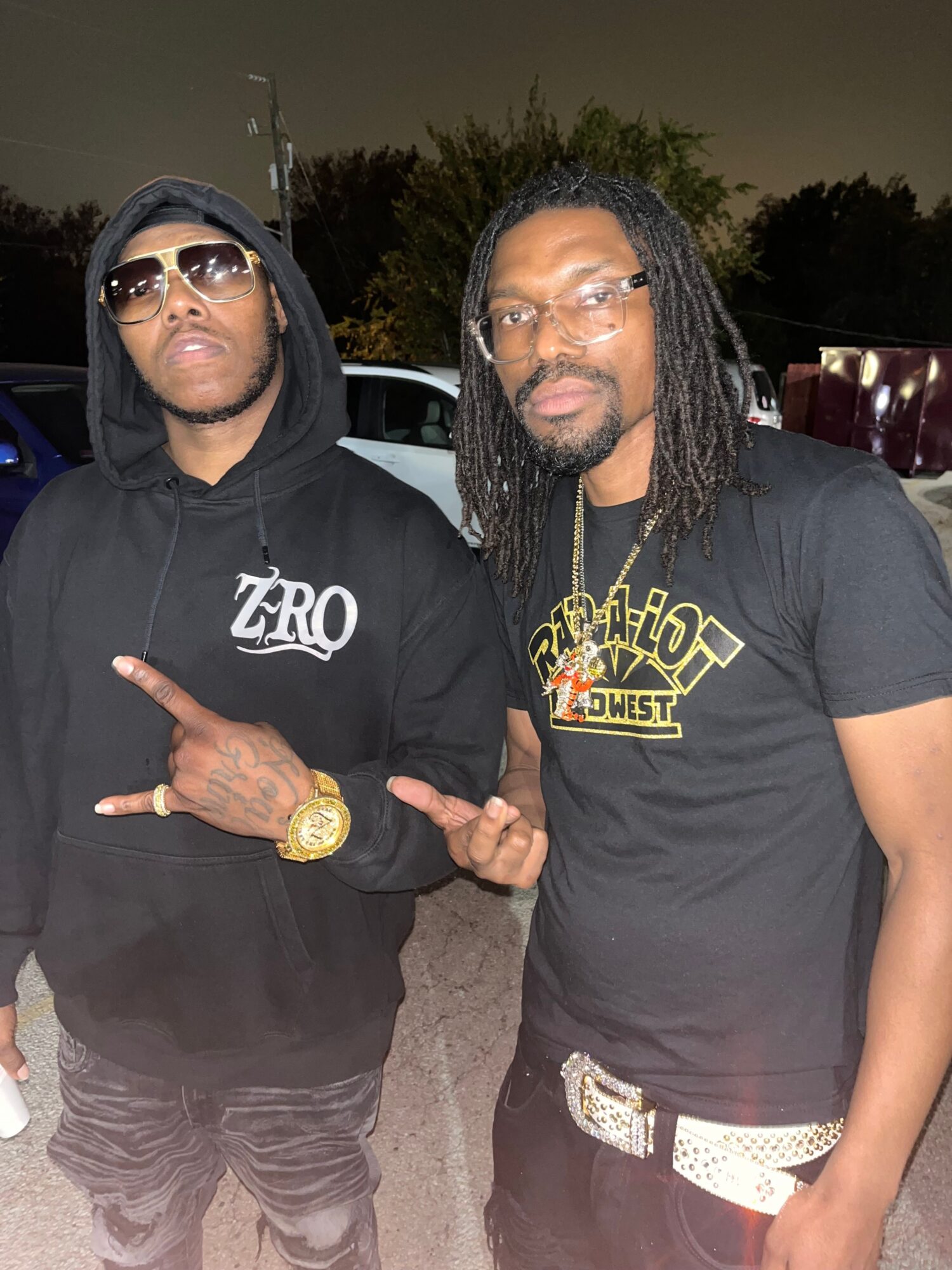 Two men standing outdoors at night, one wearing sunglasses and a hoodie, the other with dreadlocks and a T-shirt, posing together.