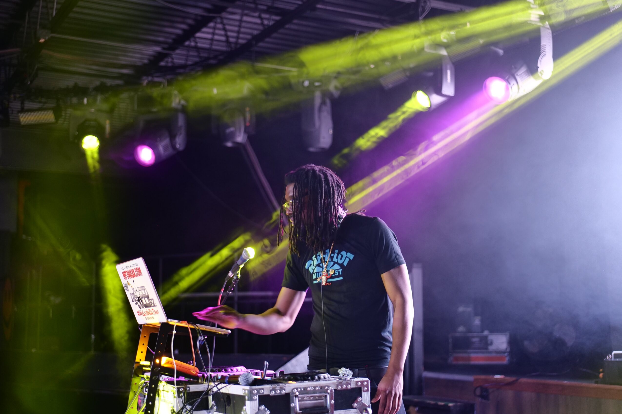 Person with dreadlocks operating electronic music equipment on stage with colorful lights and fog, in a concert setting.