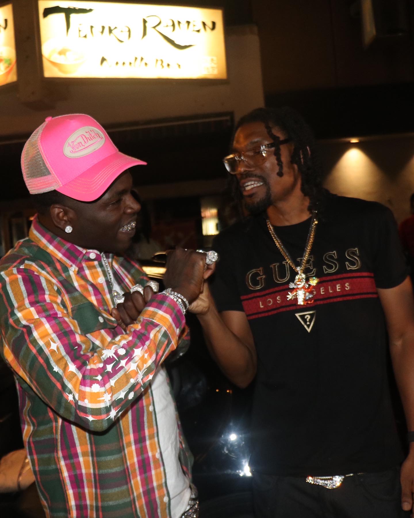 Two men smiling and fist bumping in a dimly lit indoor setting, one wearing a colorful plaid shirt and pink cap, the other in a black T-shirt with glasses.
