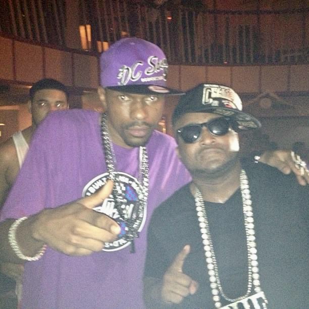 Two men pose together, one wearing a purple cap and shirt, the other in sunglasses and a black cap, both making hand gestures.