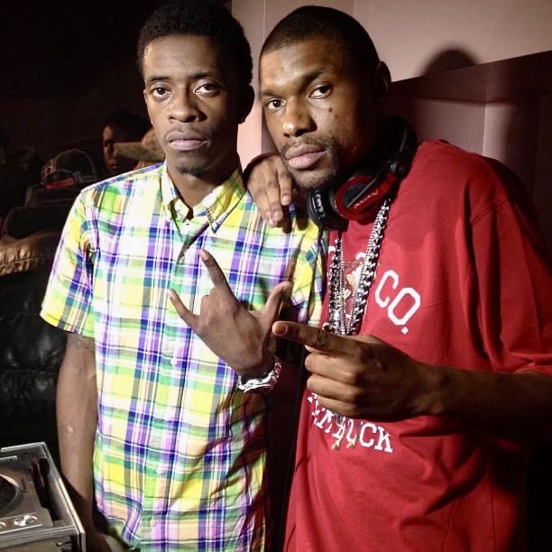 Two men pose together, one wearing a colorful plaid shirt and the other in a red shirt with headphones, making hand gestures.