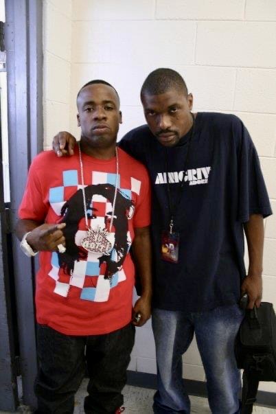 Two young men standing side by side indoors, one in a red graphic t-shirt and the other in a black t-shirt, both looking at the camera.