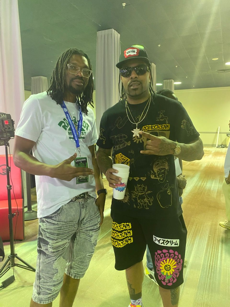 Two men standing indoors, one holding a drink and the other making a peace sign, with a green wall and window behind.