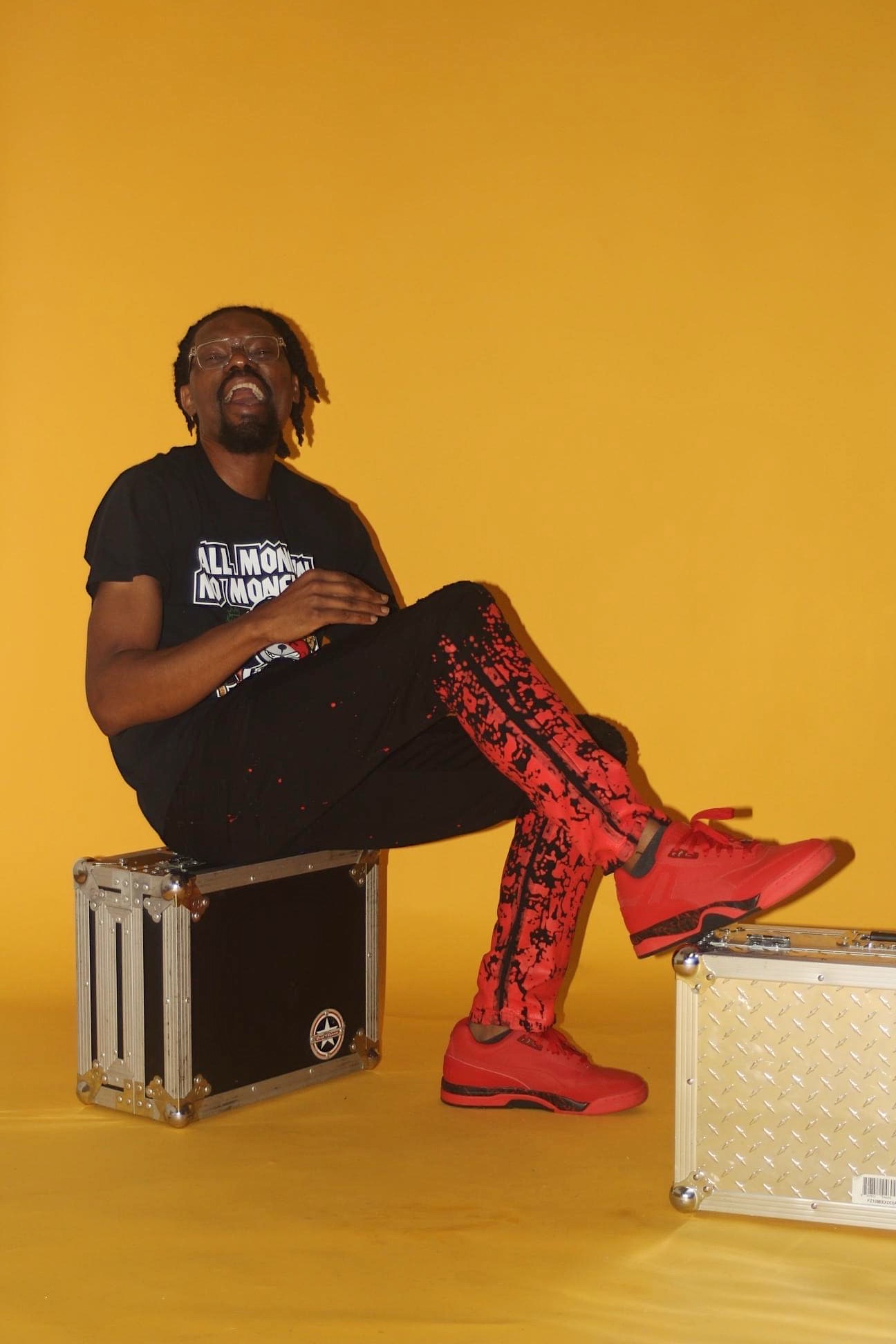 Person with dreadlocks sitting on a black case, laughing, wearing a black T-shirt and red patterned pants, against a yellow background.