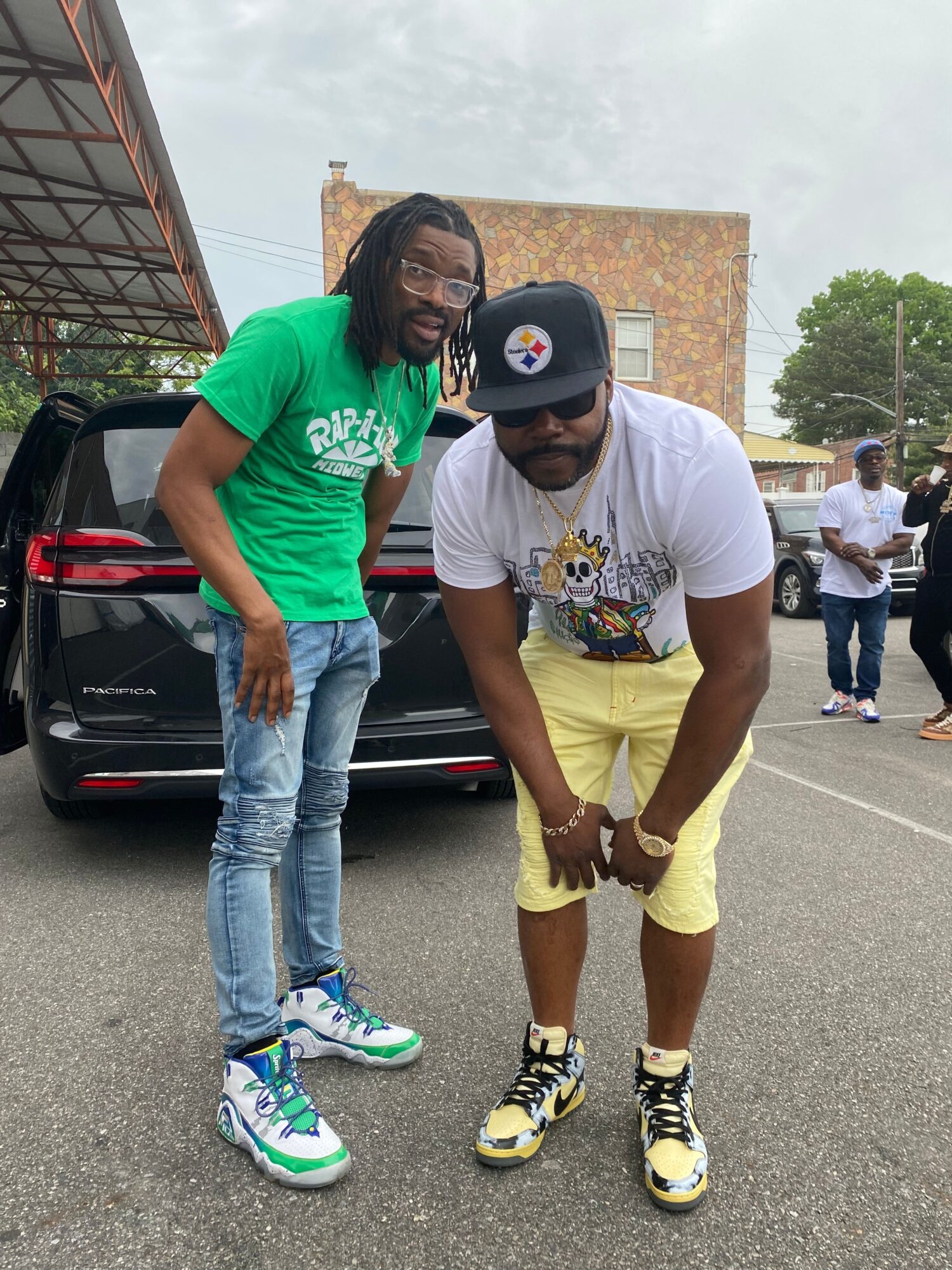Two men standing next to a black car, one in a green shirt and jeans, the other in a white shirt and yellow shorts, outdoors.